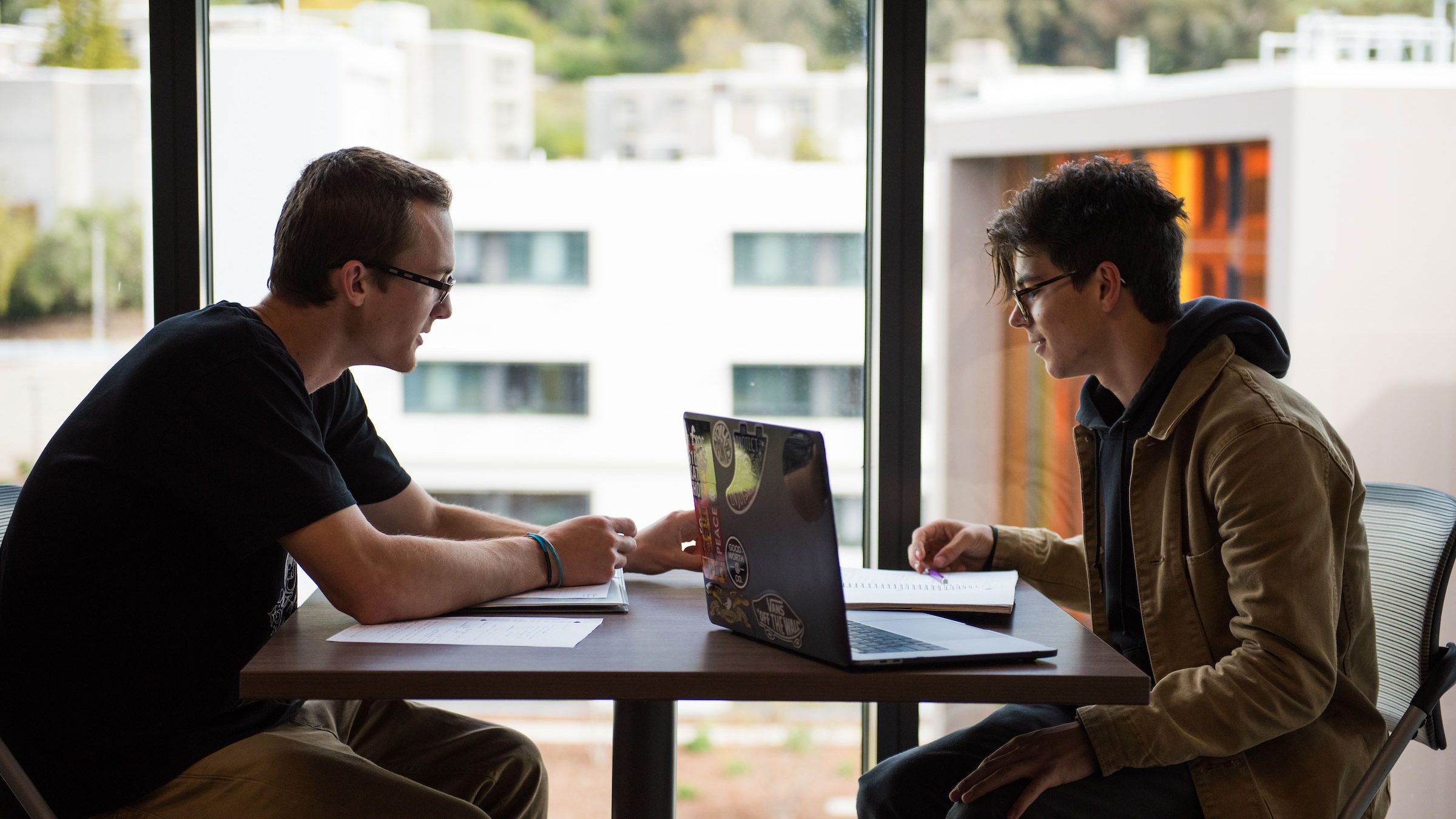 two male students facing each other at a table with laptop and notebooks